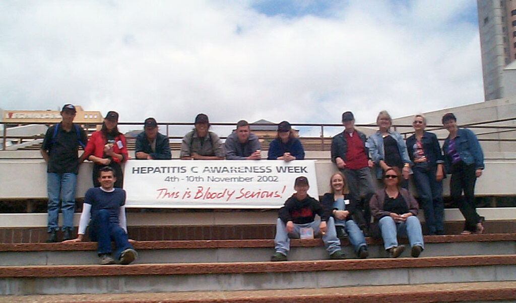Hepatitis C Council SA staff and volunteers sitting on steps around a banner that reads "Hepatitis C Awareness Week, 4th - 10 November 2002. This is Bloody Serious!"